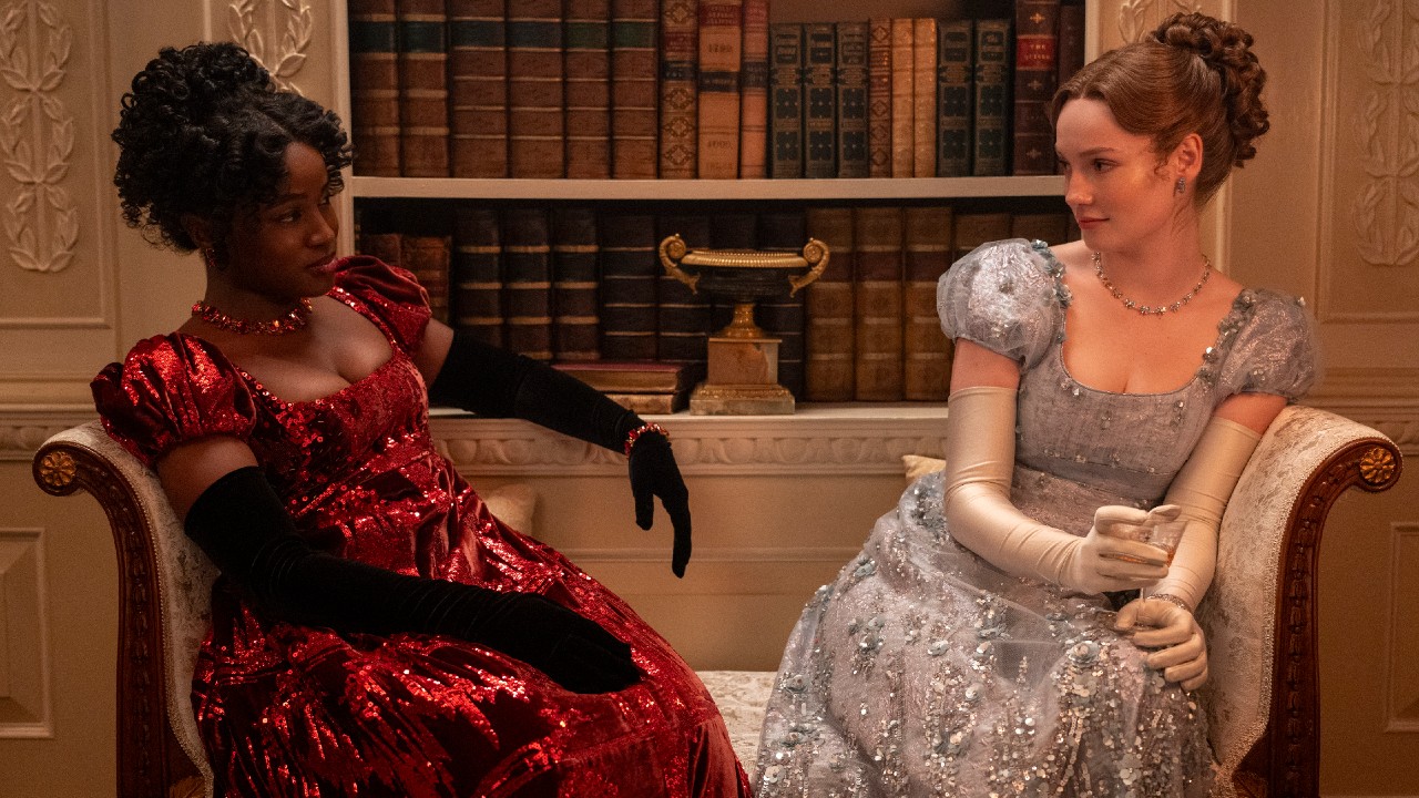 L to R) Masali Baduza as Michaela, Hannah Dodd as Francesca Bridgerton sitting on a couch and looking at each other. 