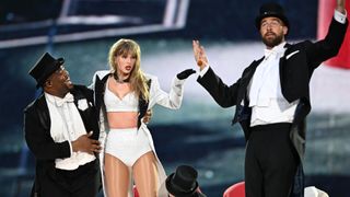 Taylor Swift wearing a white bra and briefs with a coat on stage being held up by a man in a tuxedo standing next to Travis Kelce