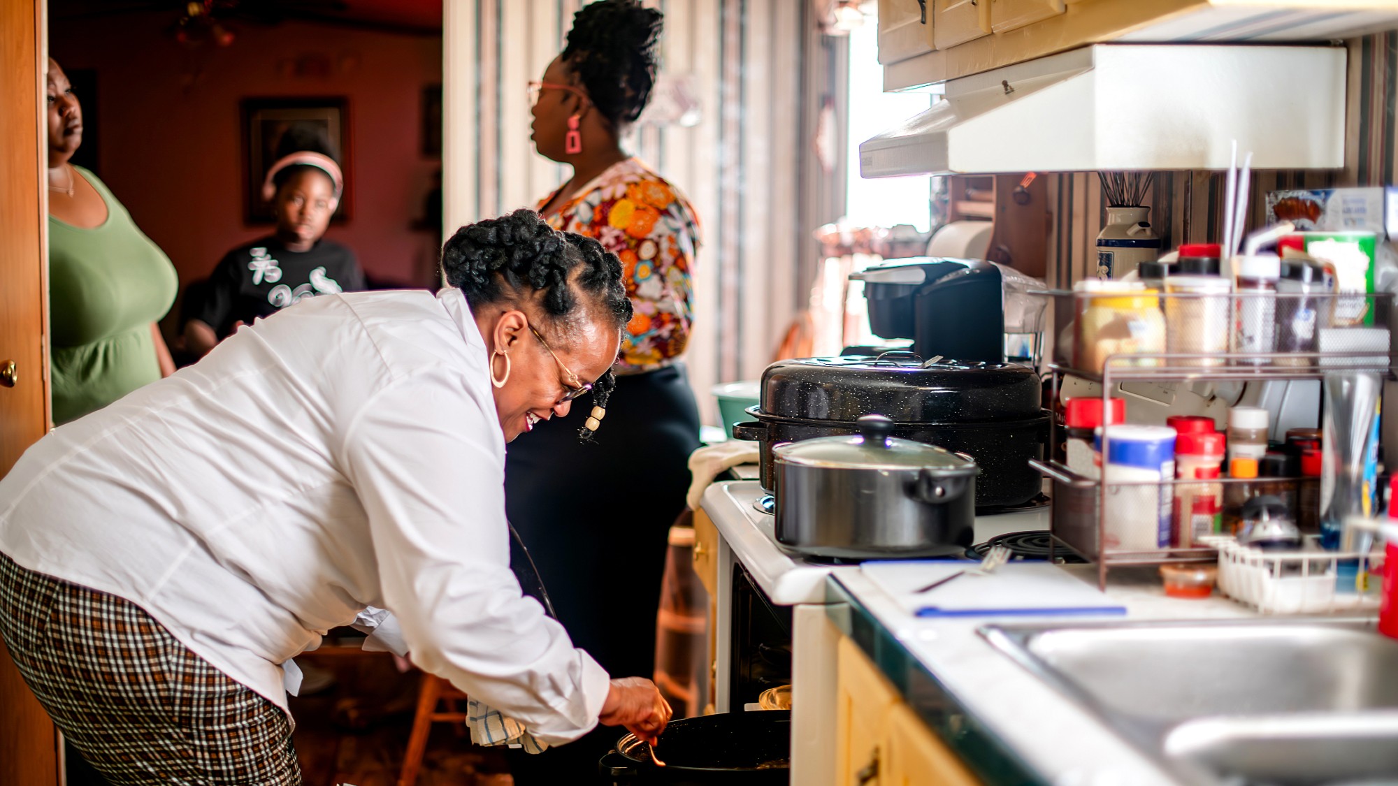 a black woman with her hair tied in braids in back stirs something in an open oven in her home kitchen. other family members are nearby