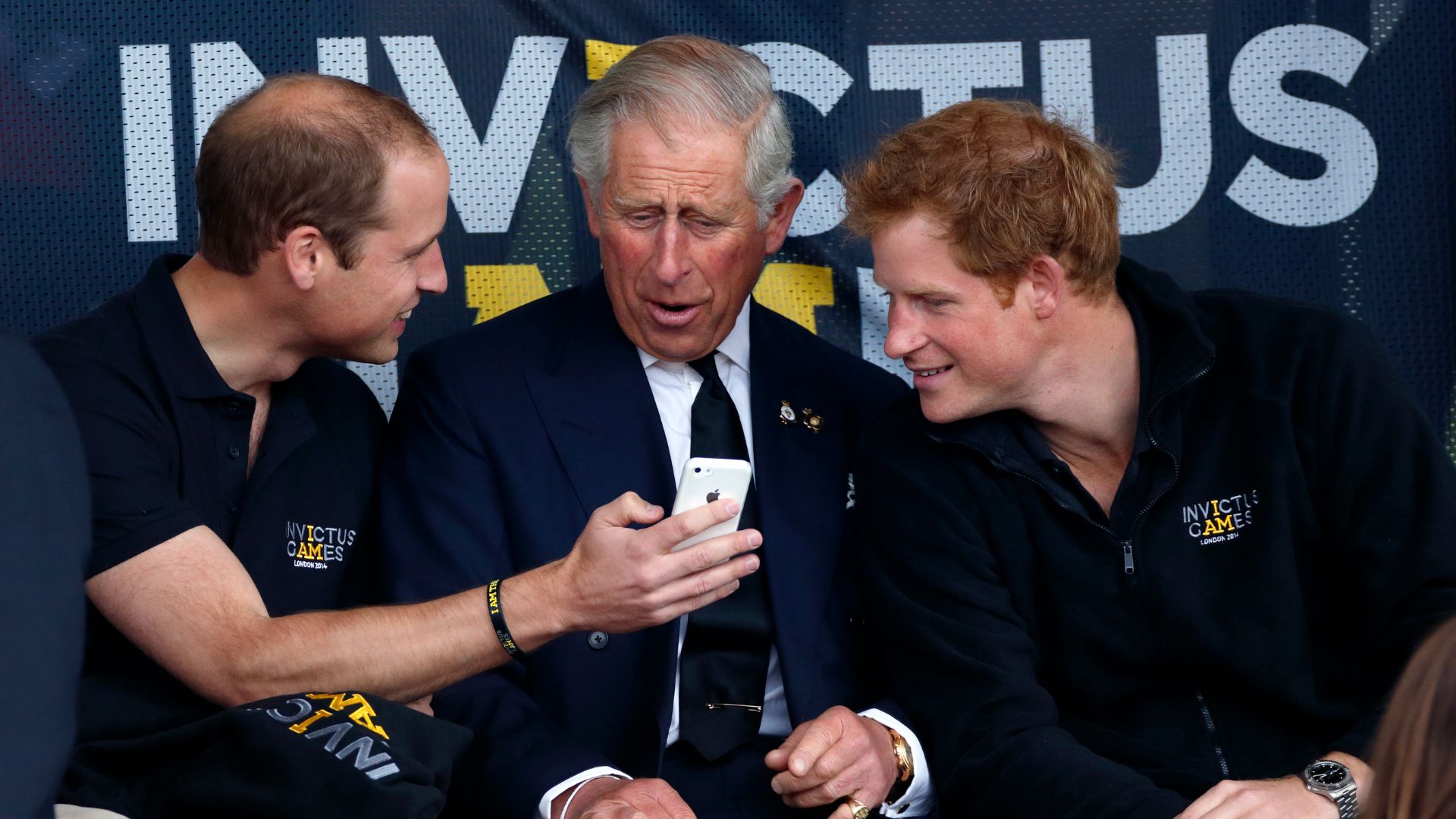 Prince William, King Charles and Prince Harry looking at a phone while attending the Invictus Games