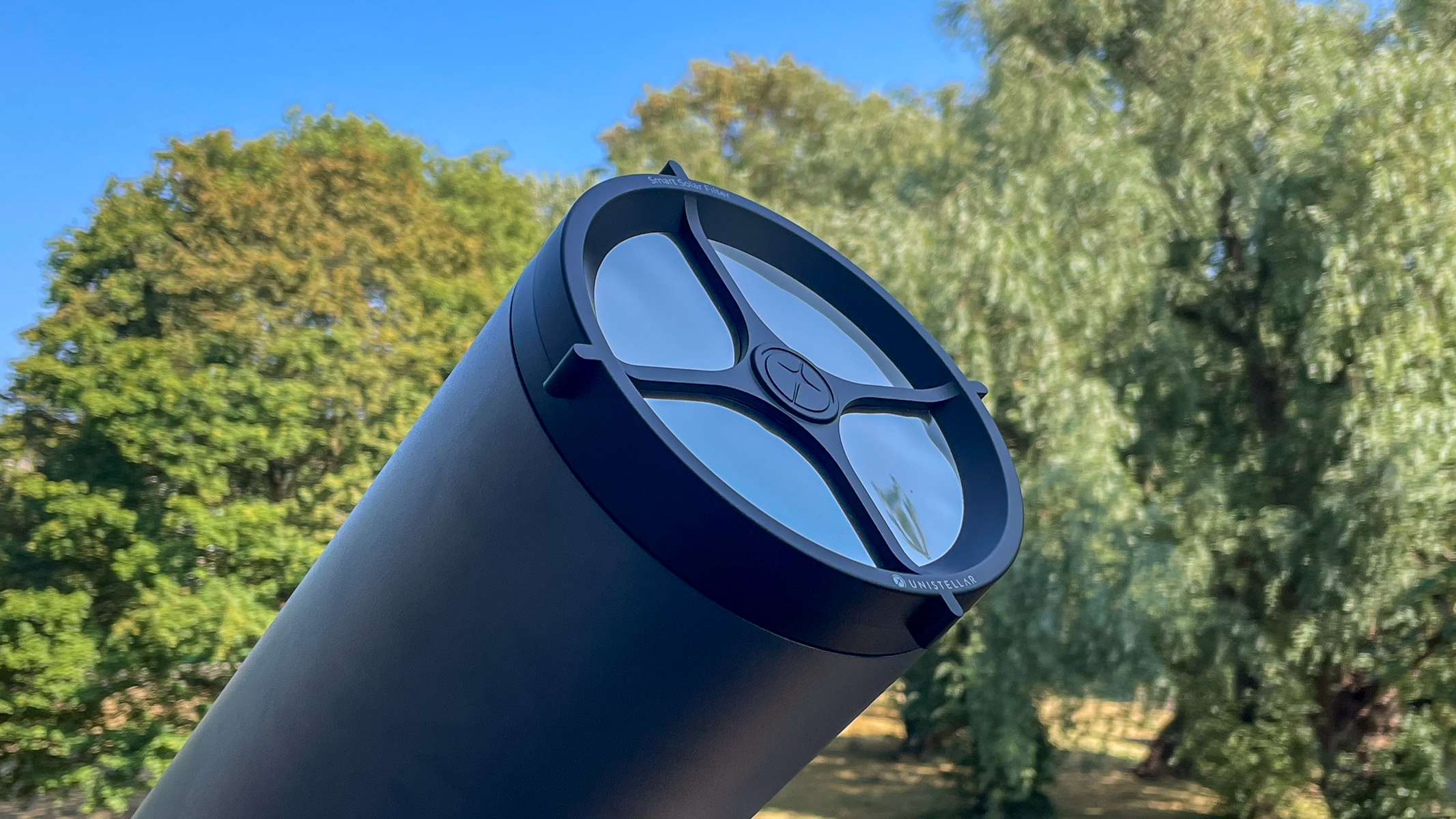 A Unistellar eVscope 2 with solar filter attached with trees in the background.