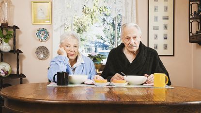 A mature couple looking regretful, sitting at a table in their robes, with breakfast in front of them.