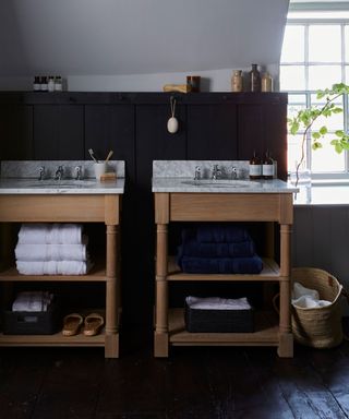 Loft bathroom with black painted walls, two wooden vanity units with marble countertops, open shelving with towels and accessories stored, black painted wooden flooring