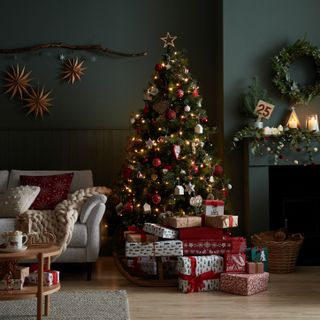 A traditional Christmas living room with tree decorated in red and green bauble decor, branch and star wall hanging, dark green wall decor, and cream upholstered sofa
