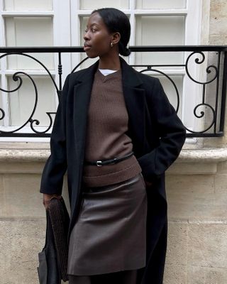 Woman wearing a brown sweater and brown skirt with a belt and long coat layered over.