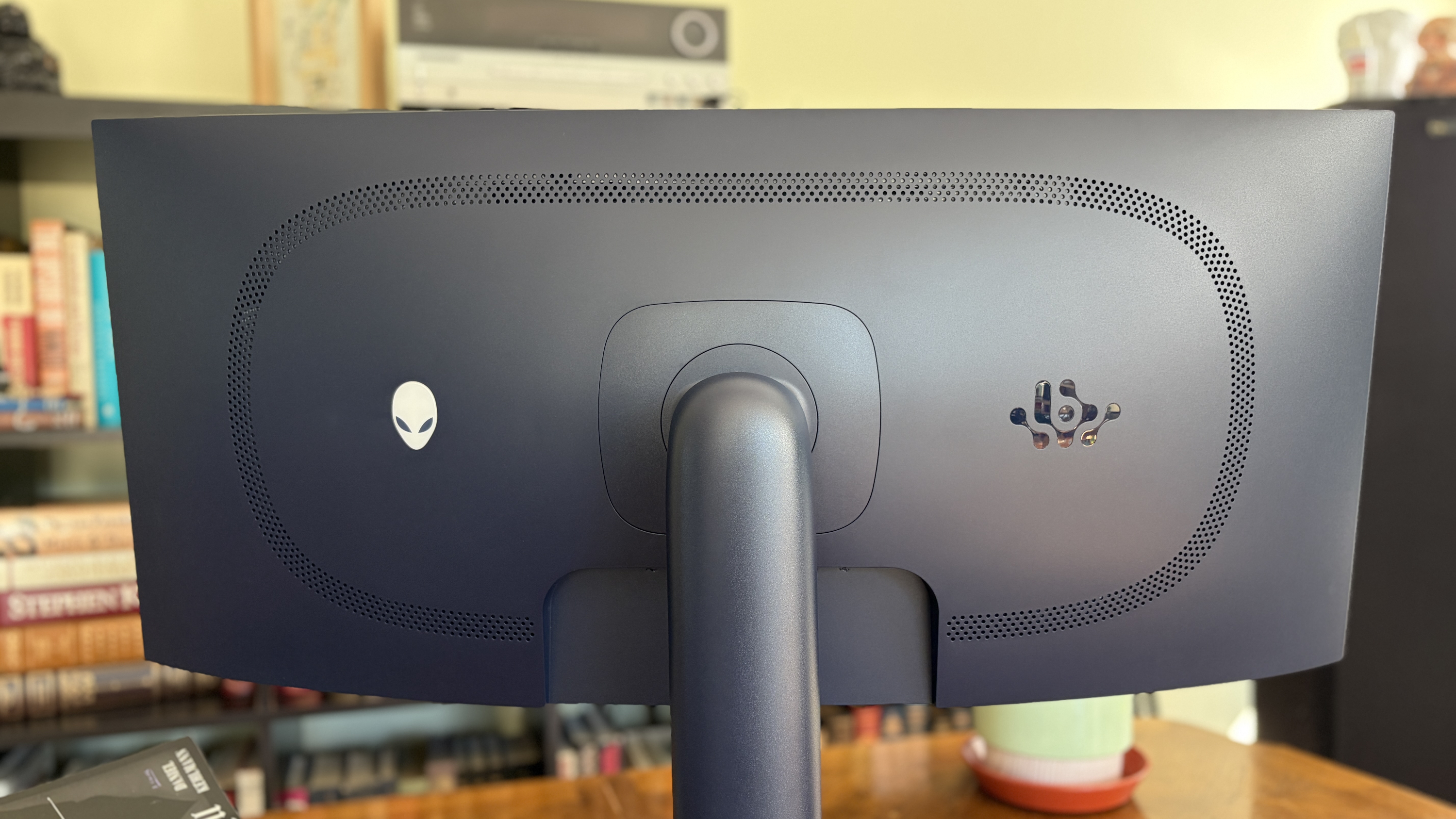 The back of a curved AW3426DW computer monitor with an Alienware logo and another emblem, with books and a potted plant in the background.