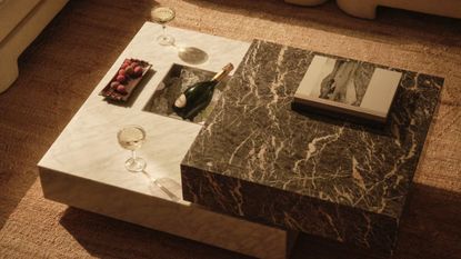 a stacked marble coffee table with built-in ice bucket with a bottle of champagne, two champagne glasses and a tray of sweets on a jute rug