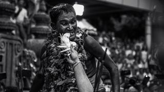 A man adorned with a leafy garland stands in a crowd, while another hand offers a ceremonial object in form of a bird during a cultural festival