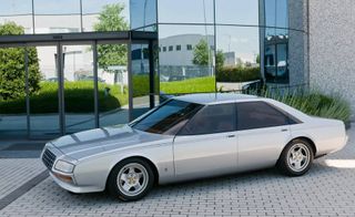 Daytime outside image of a silver 1980 Ferrari Pinin Concept, stone tiled pathway, glass fronted building, shrubs, stone wall