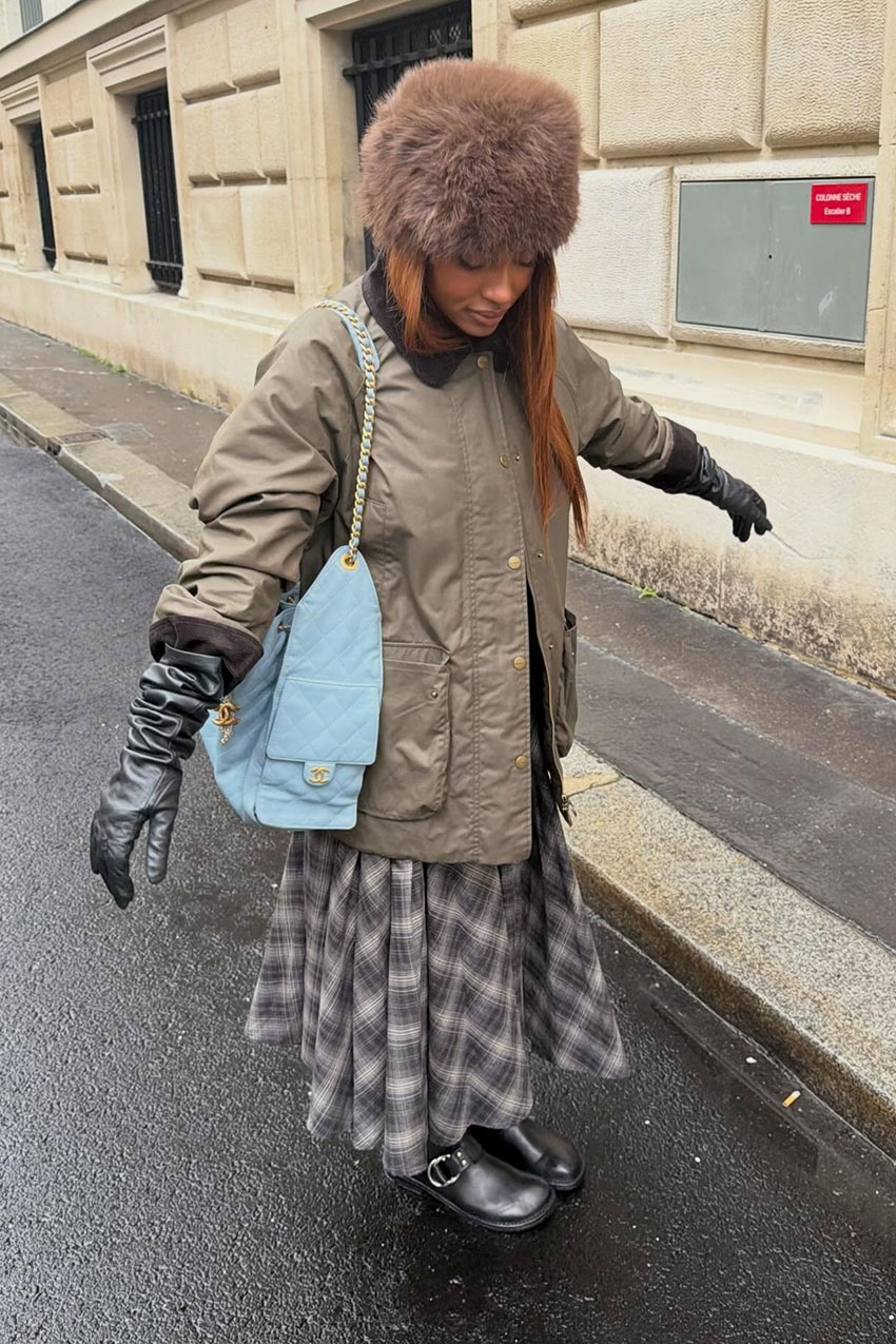 A woman wearing a fur hat, brown jacket, plaid skirt, and black leather gloves