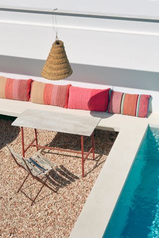 Corner of a patio in Menorca with pool, concrete bench seating, pink and orange cushions, rattan lampshade and small concrete table with red frame