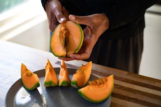 Close up image of a person's hands slicing a wedge of cantaloupe above a counter. The rest of the cantaloupe sits directly below the person's hands, already sliced into wedges. The seeds remain in the middle of each wedge.