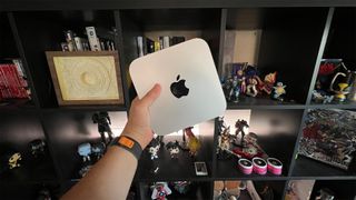 The Mac Mini Pro M2 is mini pc. It's a thin, square shape with rounded corners (20 x 20 x 3.5 cm). It's silver in color and has the Apple logo in the center of it (a black apple with a bite taken out of it). It's behind held in an adult male's hand in front of some shelves filled with video games and lots of mini anime statues.