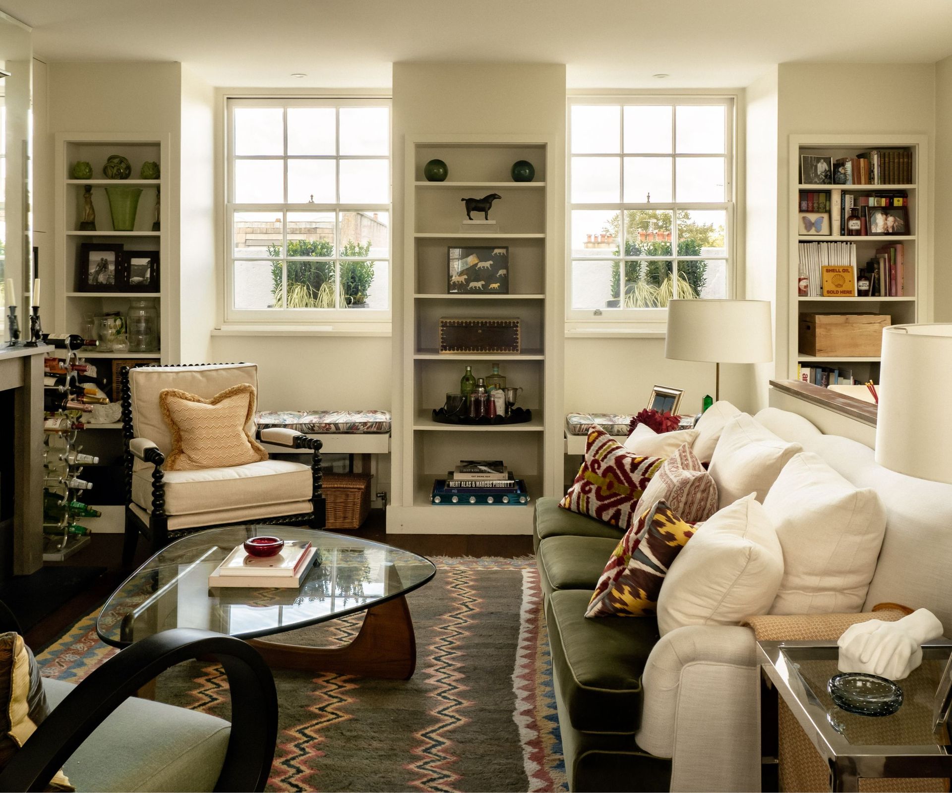 Cozy, brightly lit living room with a green velvet sofa, patterned accent pillows, a glass coffee table, built-in shelving, and large sash windows