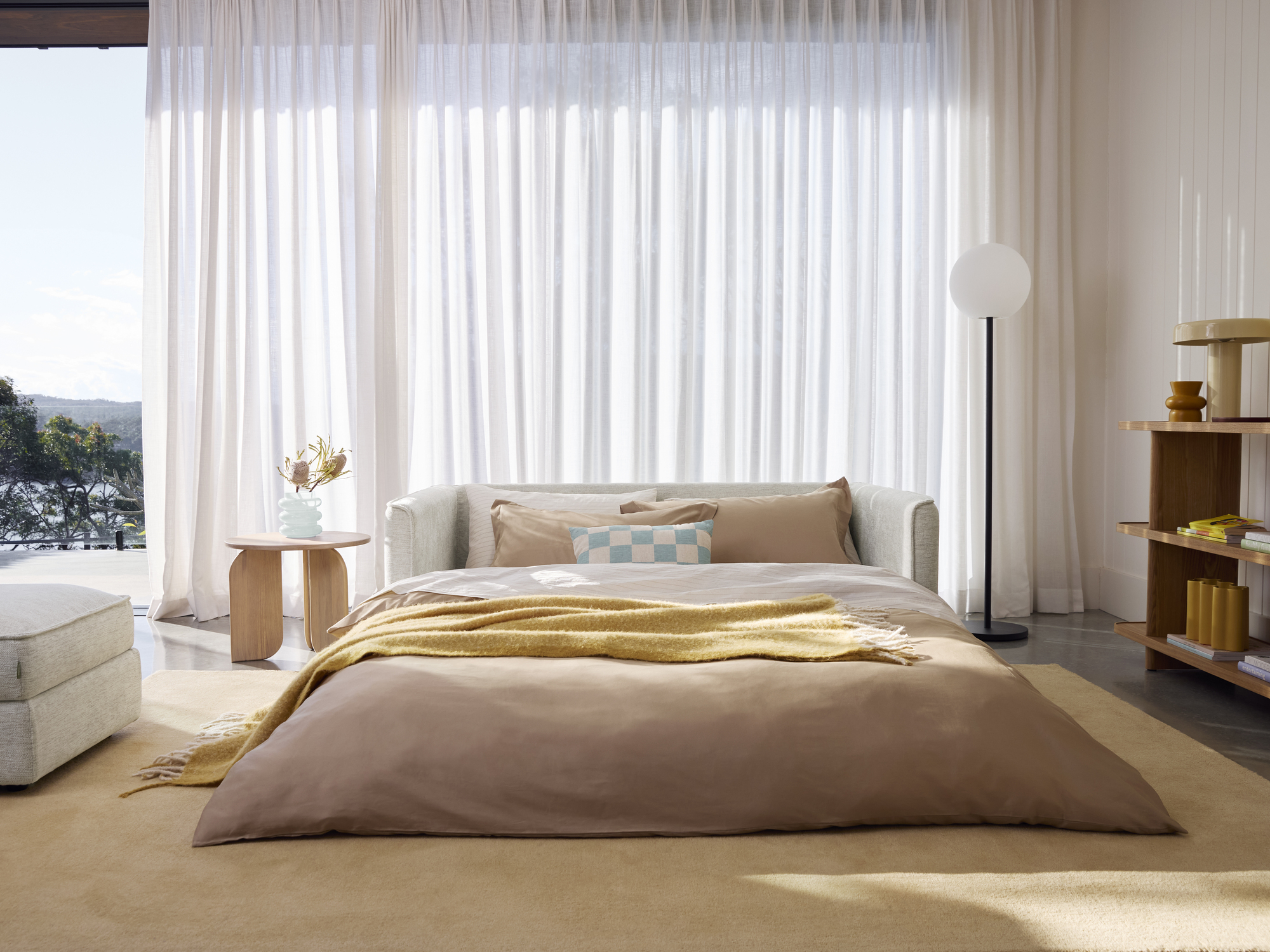 folded out sofa bed in modern room with sheer wall of curtains behind, cream rug, pink and yellow and white sheets, green checkered cushion, floor lamp, side table with vase, ottoman, and console styled with decor