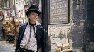 Billy Jenkins as Dodger stands in a 19th century street wearing a battered top hat, dark jacket and white shirt in Dodger.