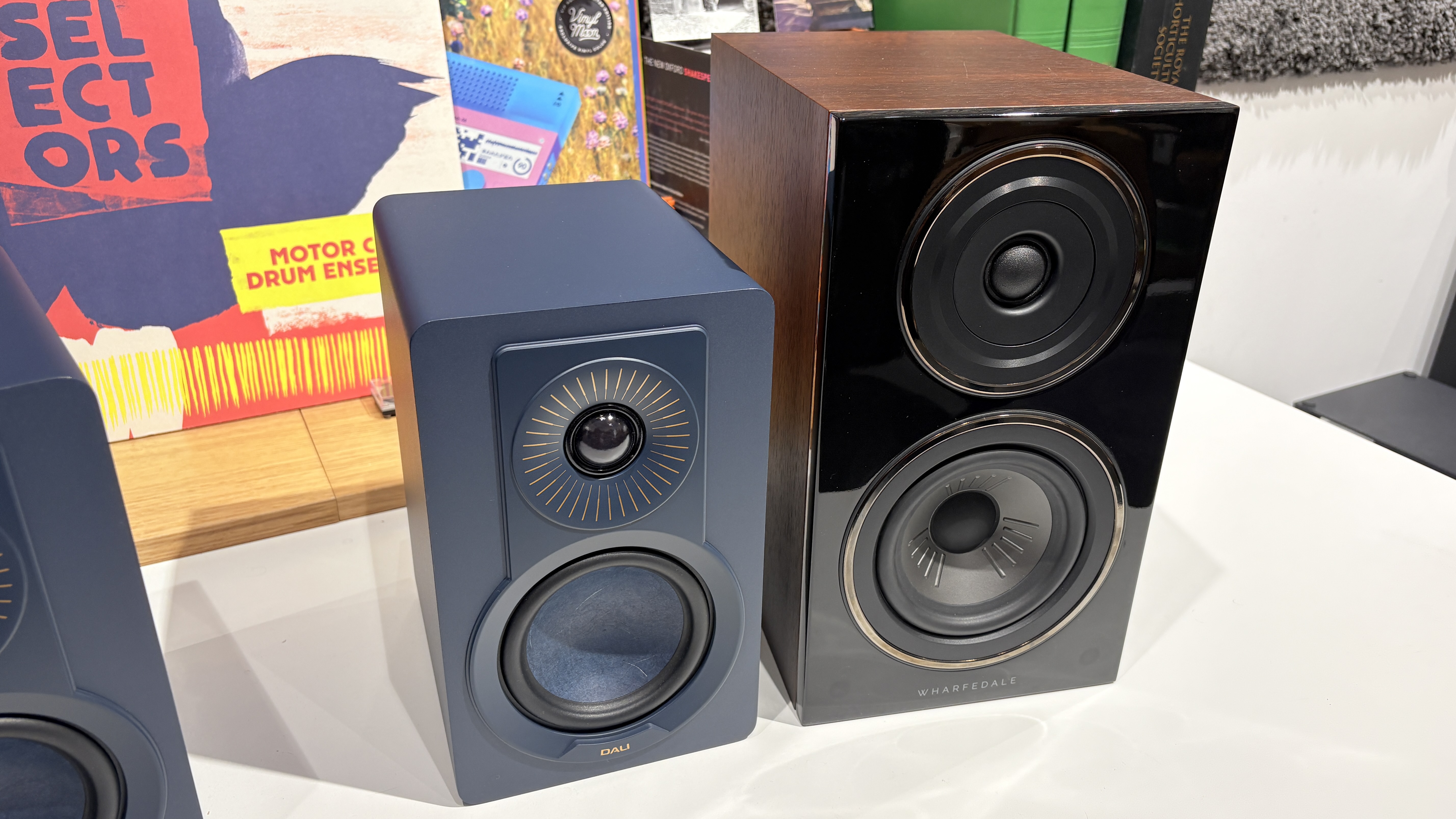 Dali Kupid speaker in blue finish next o Wharfedale Diamond 12.1 in walnut finish, both on a white desk