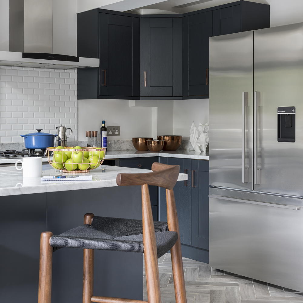 black kitchen cupboards with white marble tops with a silver stainless steel fridge and a island topped with apples in a bowl and accessorizd with a wooden chair with a black woven sitting-top