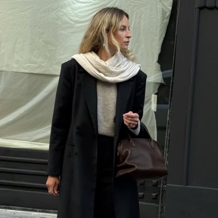 A woman wears a black coat and pants with a beige sweater, cream scarf, and leather boots.