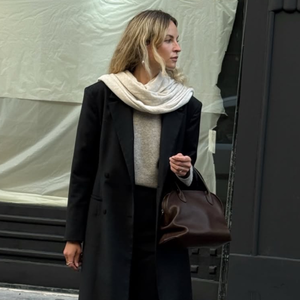 A woman wears a black coat and pants with a beige sweater, cream scarf, and leather boots.