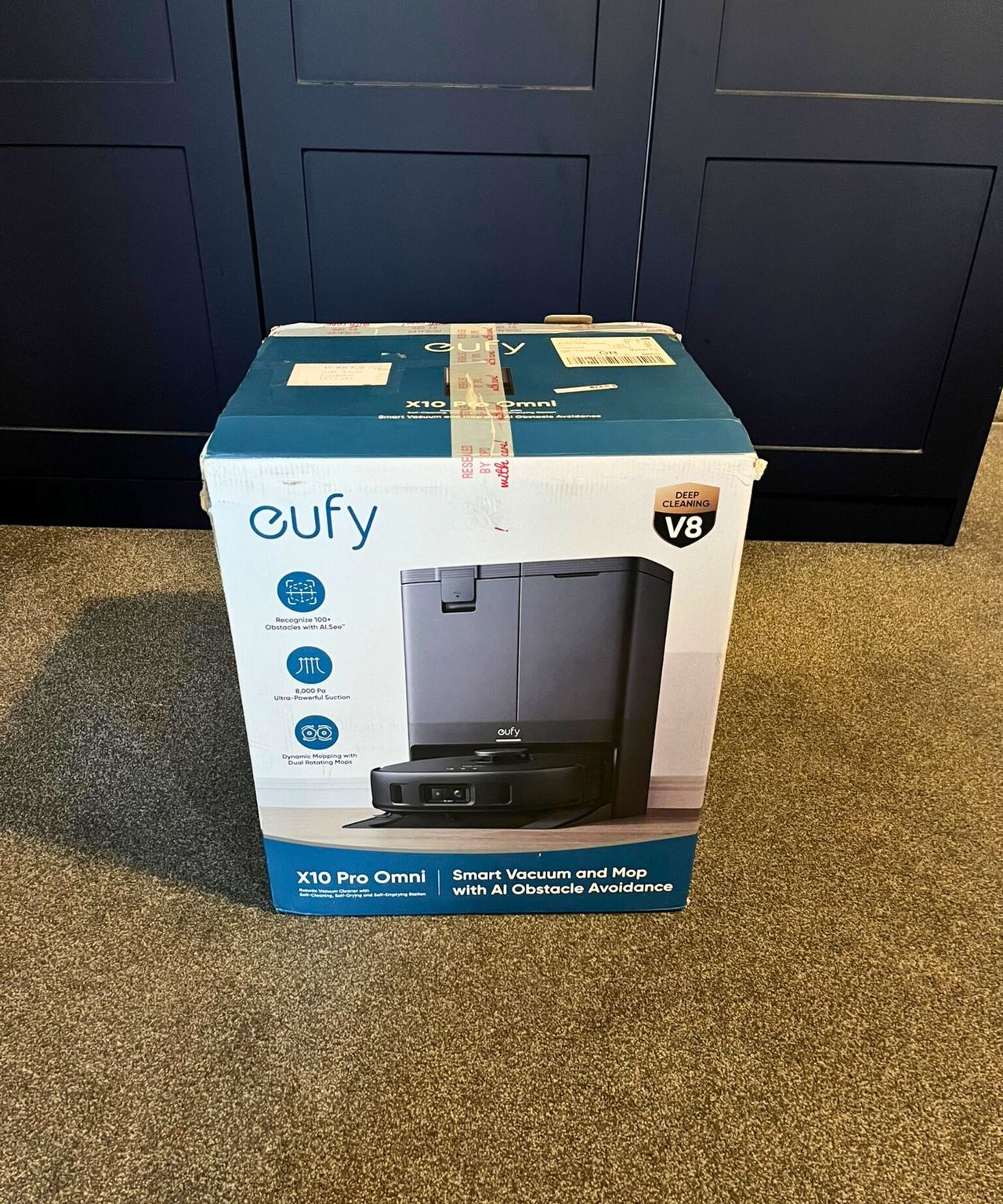 eufy X10 Pro Omni robot vacuum in its delivery box which carries an image of the item. It's on a gray carpet in a bedroom with fitted matte navy blue closets
