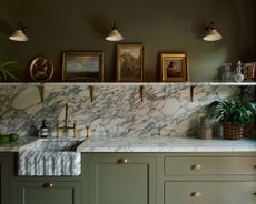 A Kitchen with a marble worktop, marble sink and marble splashback. There is a marble shelf above the splashback with four paintings in a gold frame. The cupboards are a deep mossy brown colour. 