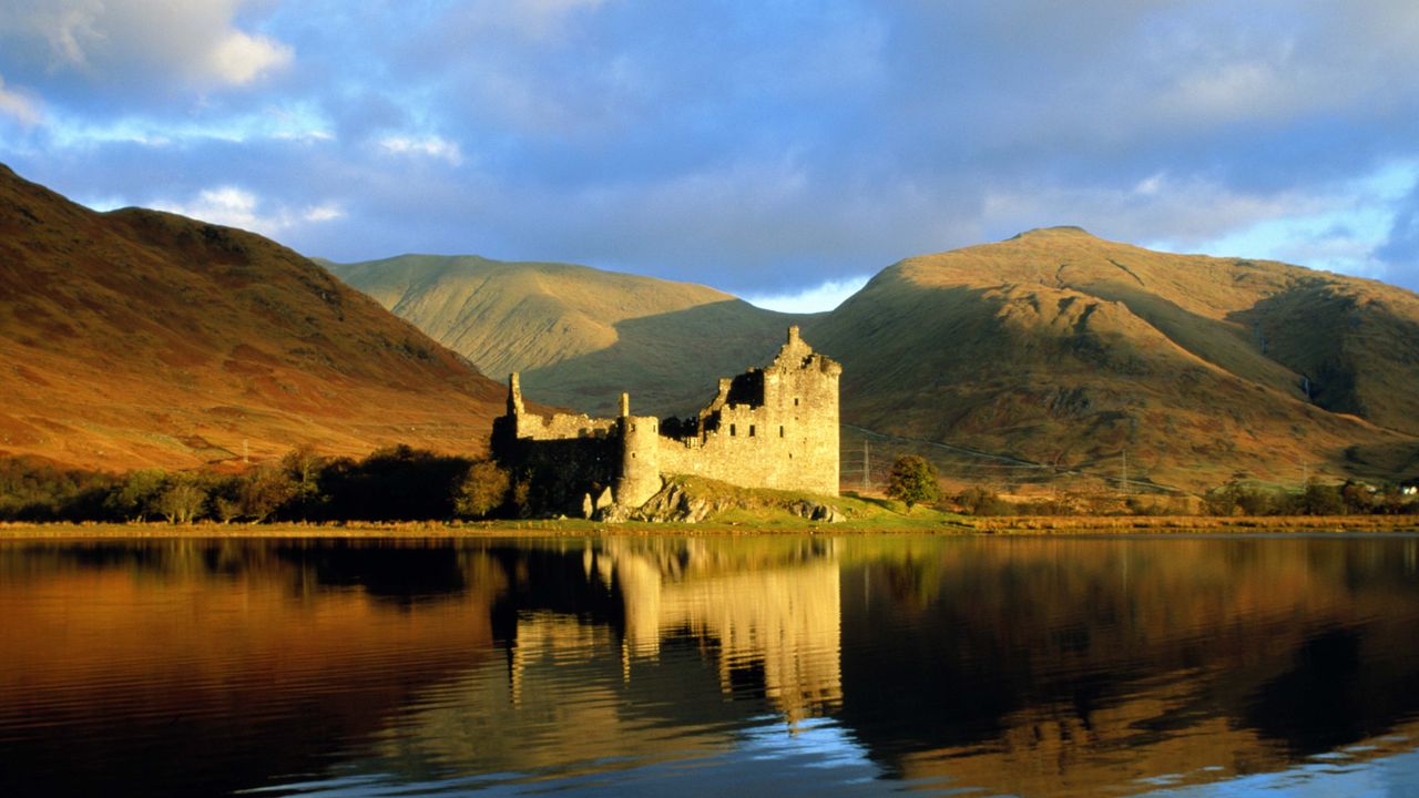 A photo of one of the castles in Scotland set against a Loch with mountains behind 