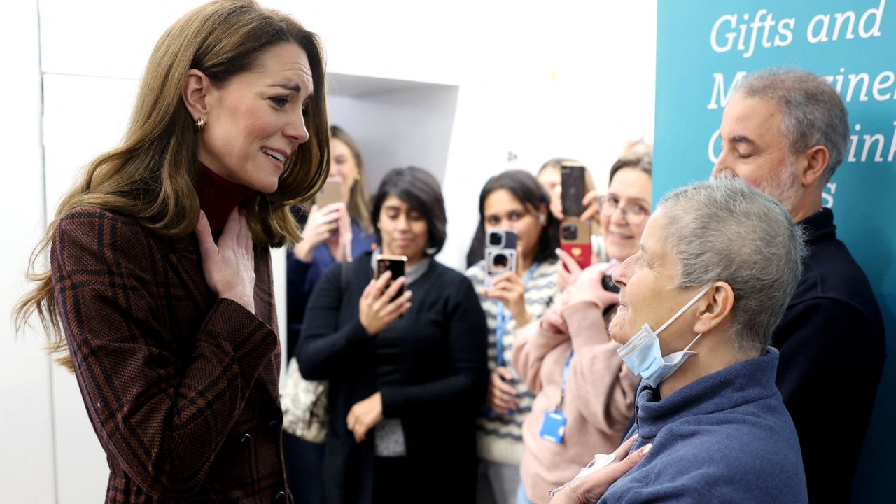 Kate Middleton wearing a plaid coat looking emotional and putting her hand against her chest talking to a woman with a mask pulled down as people take photos in the background