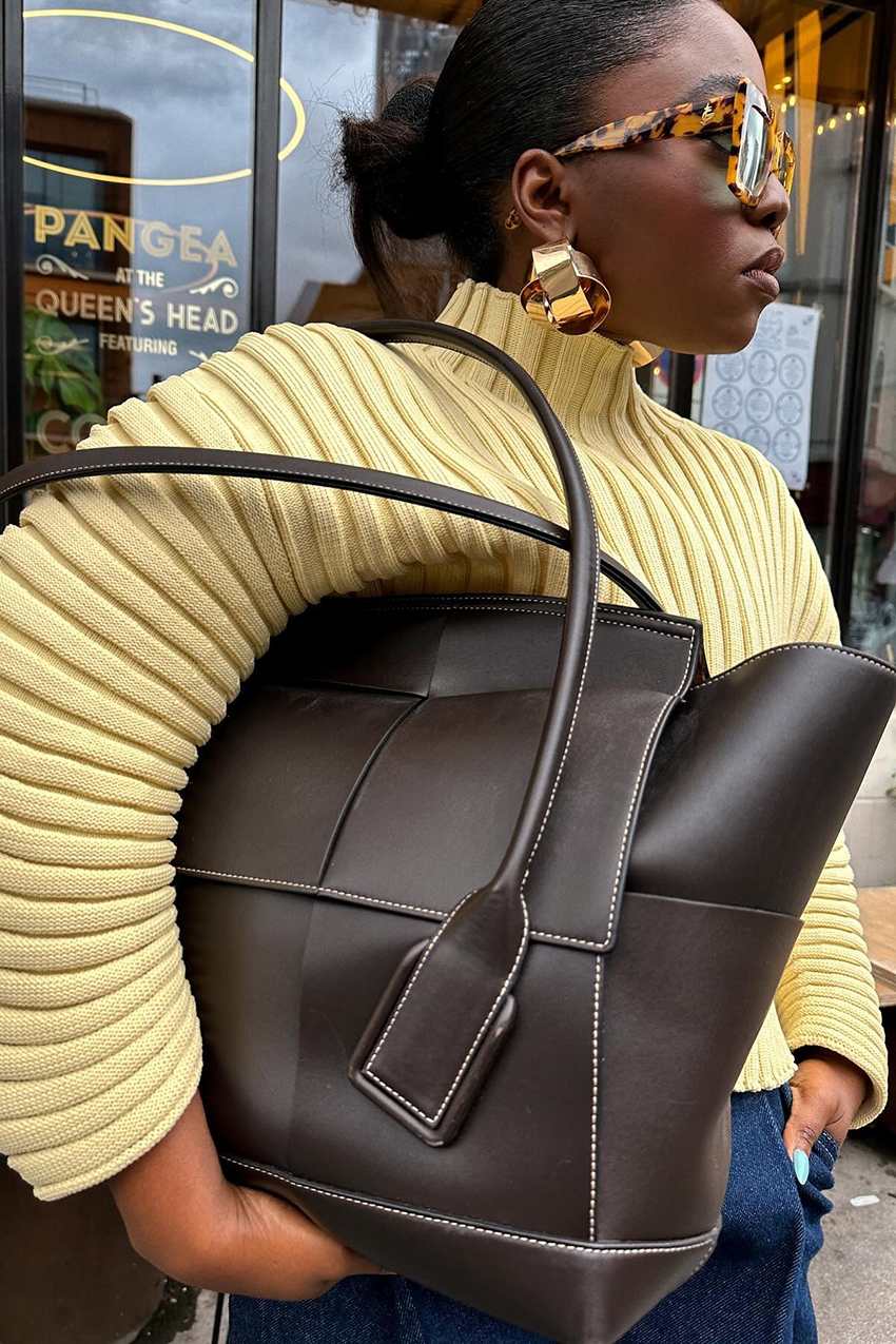 a woman wearing a butter yellow sweater, brown leather bag, tortoise shell sunglasses, and gold earrings