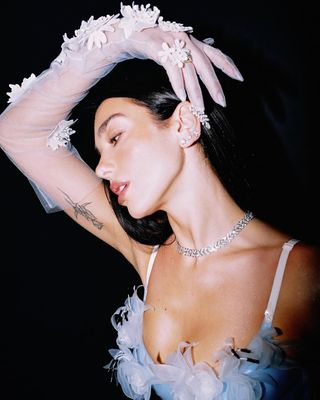 A photo of Dua Lipa accessorizing her translucent dress with a matching pair of organza opera gloves.