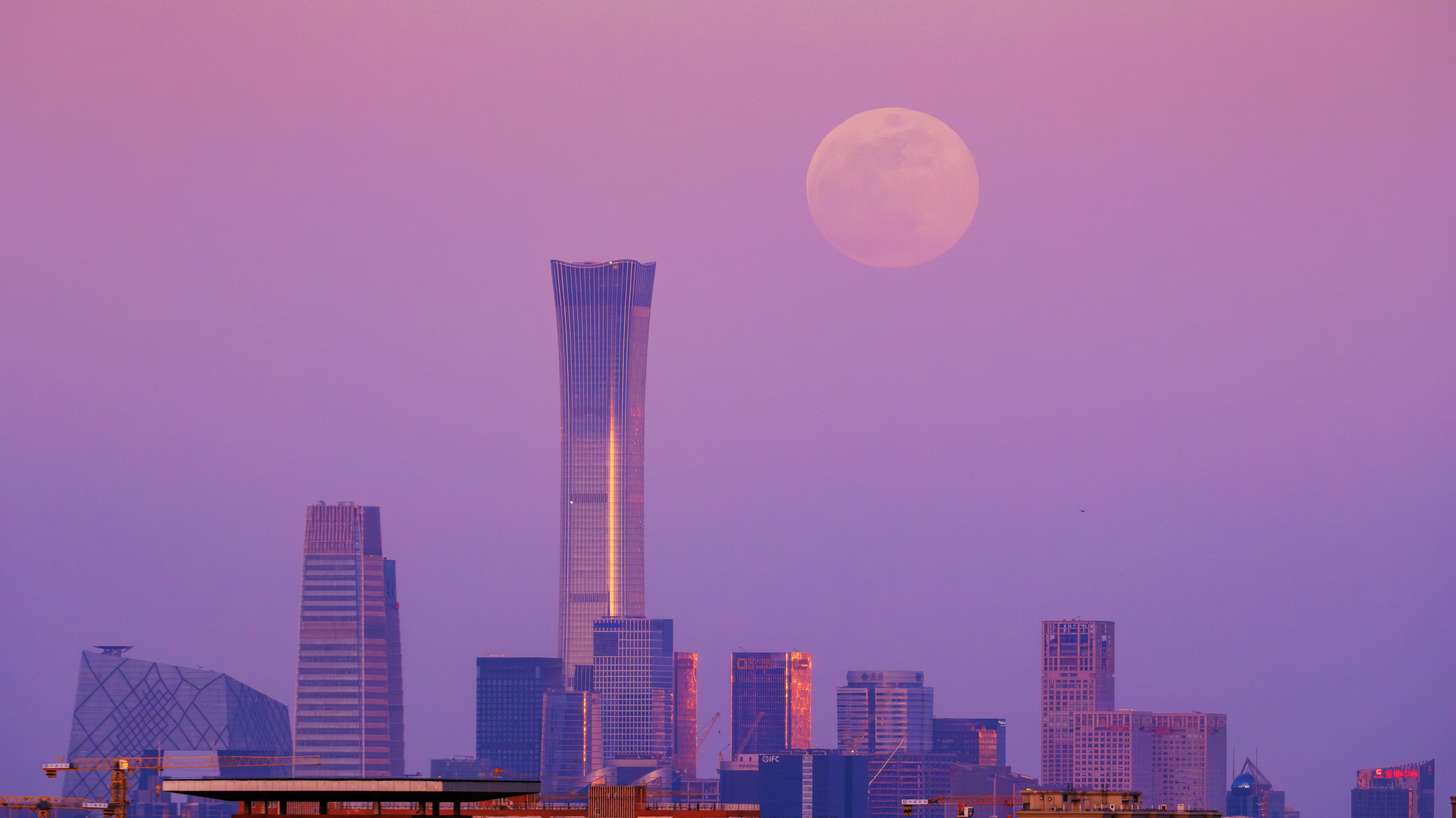 february full moon shines delicately in a pink purple sky above a city skyline with tall skyscrapers.