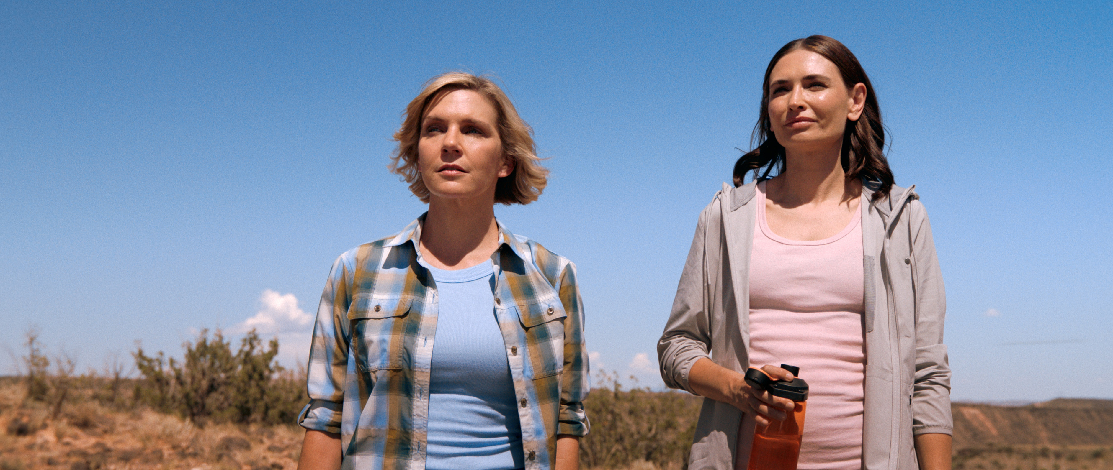 Two women (Rhea Seehorn as Carol and Karolina Wydra as Zosia) stand in the New Mexico desert on a sunny day. Carol wears a plaid shirt and Zosia wears a gray hoodie while holding a water bottle, in a still from 'Pluribus.'
