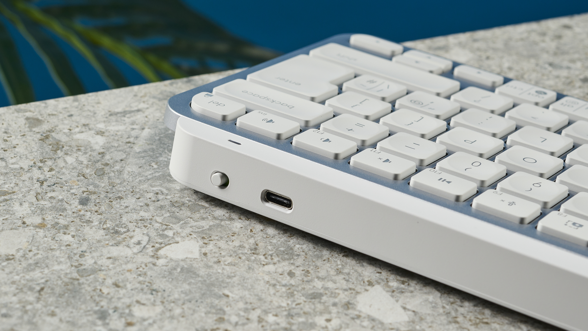 A photograph of the Logitech MX Keys Mini in light gray, positioned on a stone slab with a blue wall in the background.