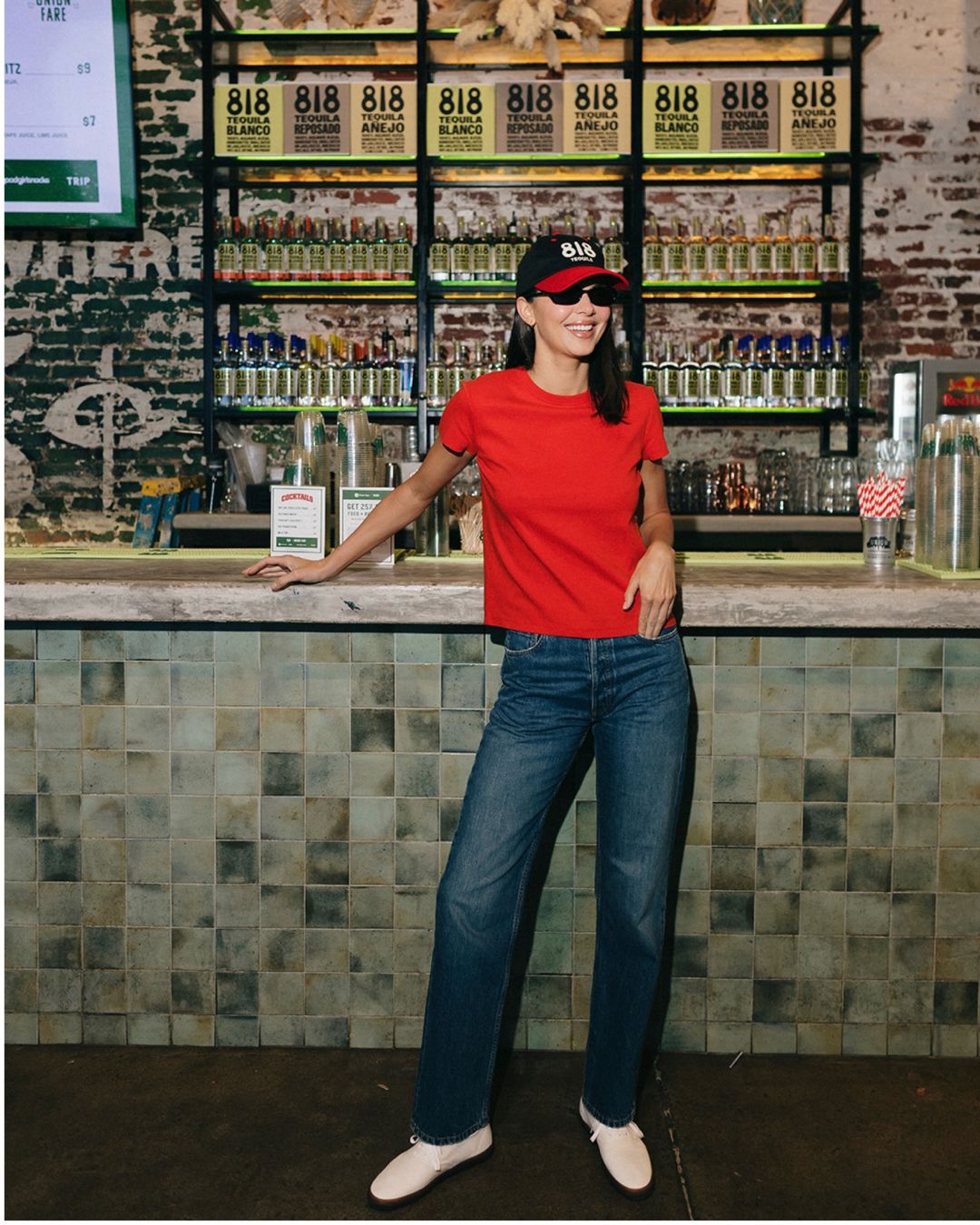 Kendall Jenner poses at a bar wearing dark wash jeans with white Derby shoes and a red t-shirt.