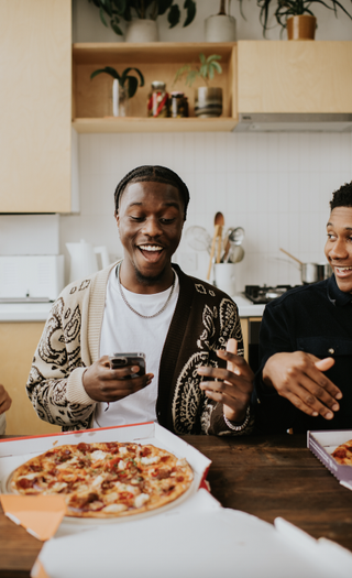 Three young people have ordered a food delivery. It has arrived in flat cardboard boxes. The friends sit at a dining table, and open the box to reveal a delicious pizza with various toppings. The vibe is relaxed, and fun.