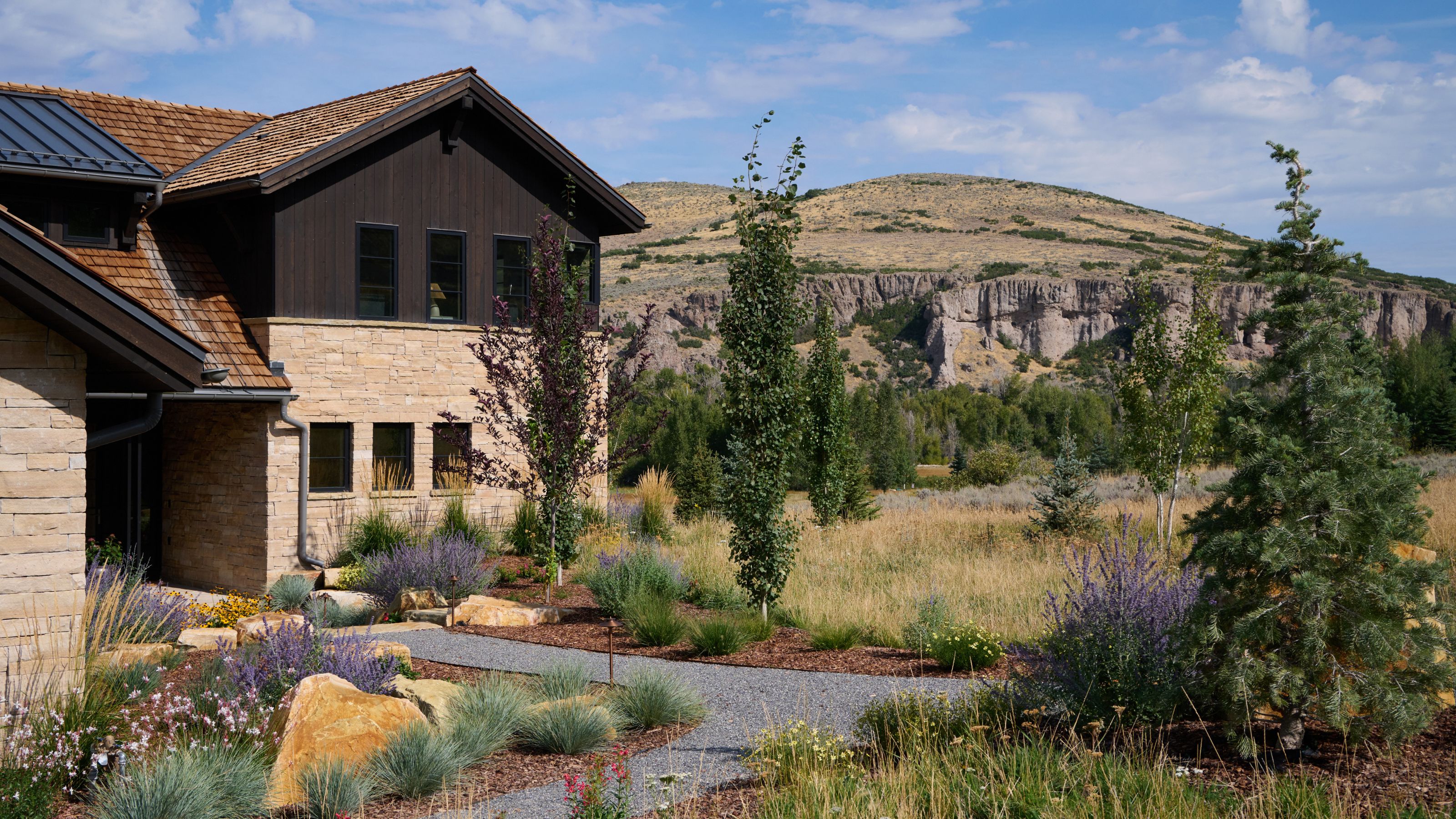 Stone and wood house exterior with landscaped garden of shrubs, flowers, and small trees, with cliffs and hills in the background.