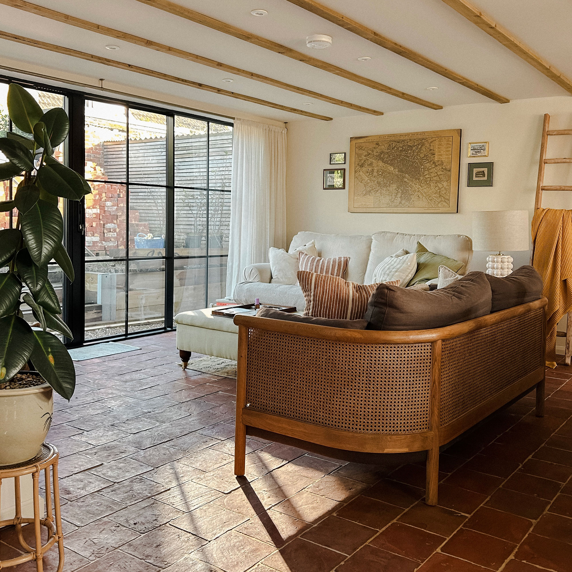 Living room with crittall style doors, terracotta tiled floor, cane backed sofa and map as artwork on wall