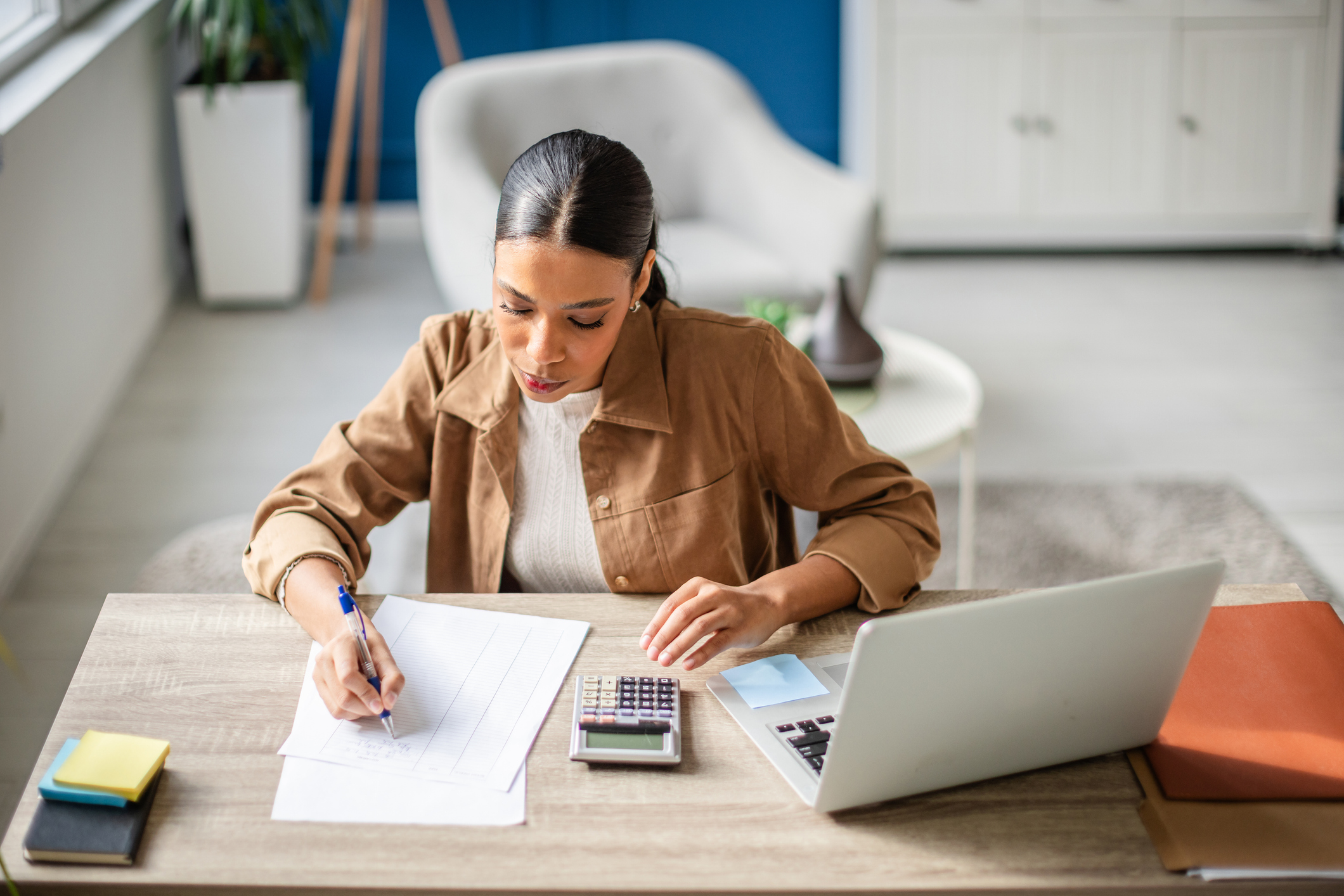 A young woman is focused on preparing her taxes at a home office. She writes notes, uses a calculator, and organizes her financial documents on the desk.