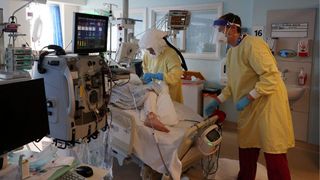 Two people in yellow gowns, gloves, face shields and masks tend to a COVID-19 patient in a hospital bed