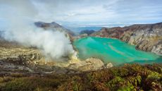 View of Kawah Ijen crater lake from the top of the volcano. The lake's waters are bright turquoise and there is a plume of gas rising to the left.