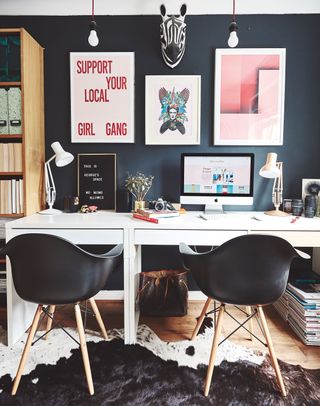 a white desk with two black chairs, with a cow-print rug, a dark blue wall, pink prints and stationary on the desk