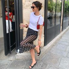 French style influencer Leasy In Paris poses outside a cafe in Paris wearing a claw hair clip, black sunglasses, yellow-gold chunky earrings, a white contrast hem T-shirt, mini red shoulder bag, bangles, a polka-dot skirt, simple silver watch, and black kitten-heel mule sandals