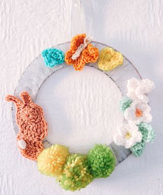 A silver ribbon wreath with colorful crocheted decorations, with a pink bunny, yellow and green easter eggs, white flowers with blue leaves, and a butterfly with yellow and blue flowers next to it