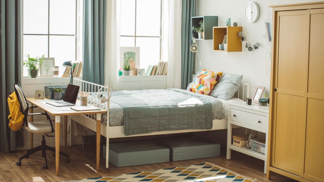 A dorm room with a metal bed, a desk at the bottom of it with a computer, under bed storage, and a yellow closet