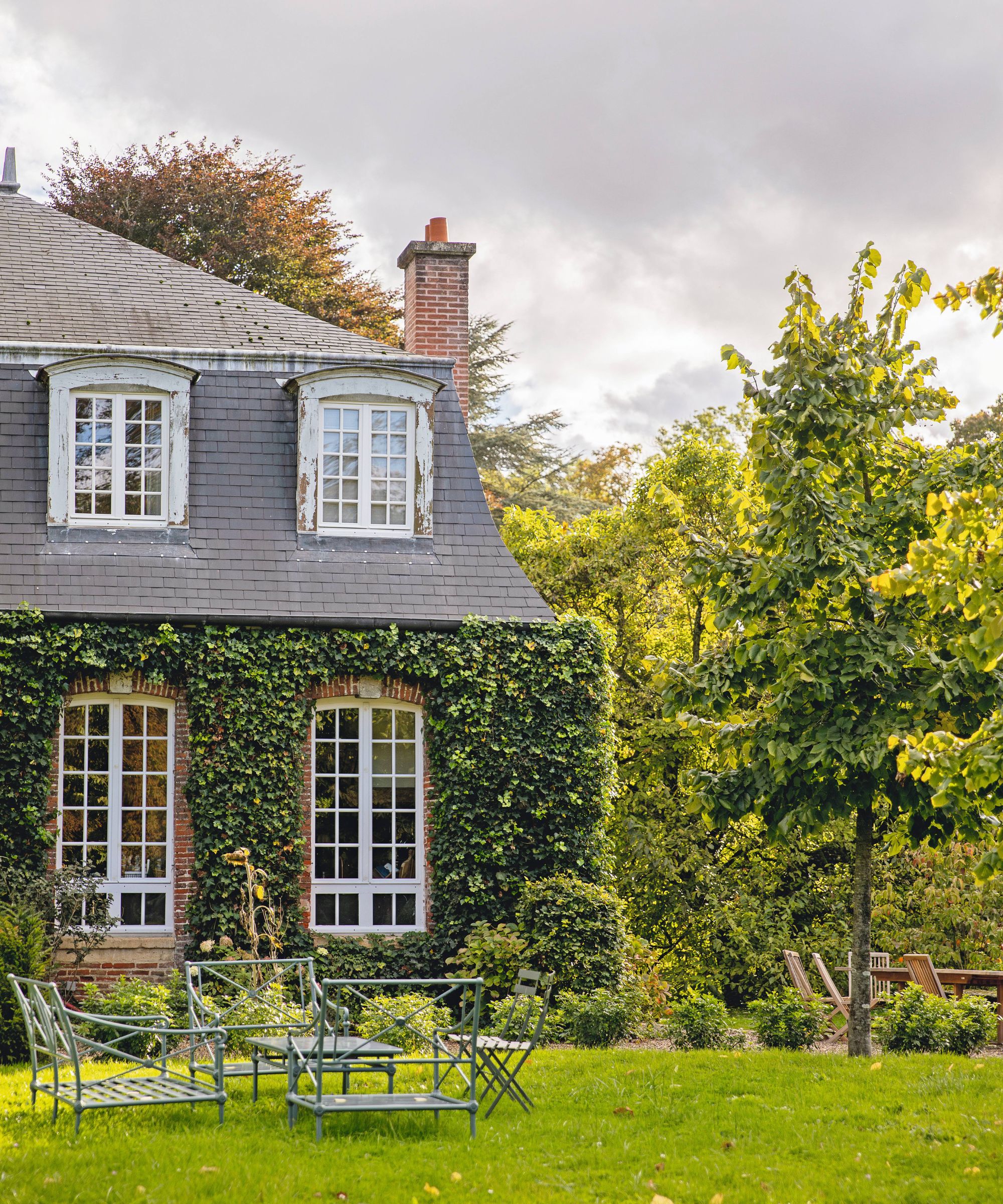 an exterior shot of a french chateau home with large windows, chimney, and a lawn area with a wrought iron garden table and chairs set