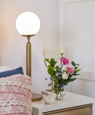 A tall brass lamp with white and pink flowers next to it, as well as throw pillows on a bed next to it