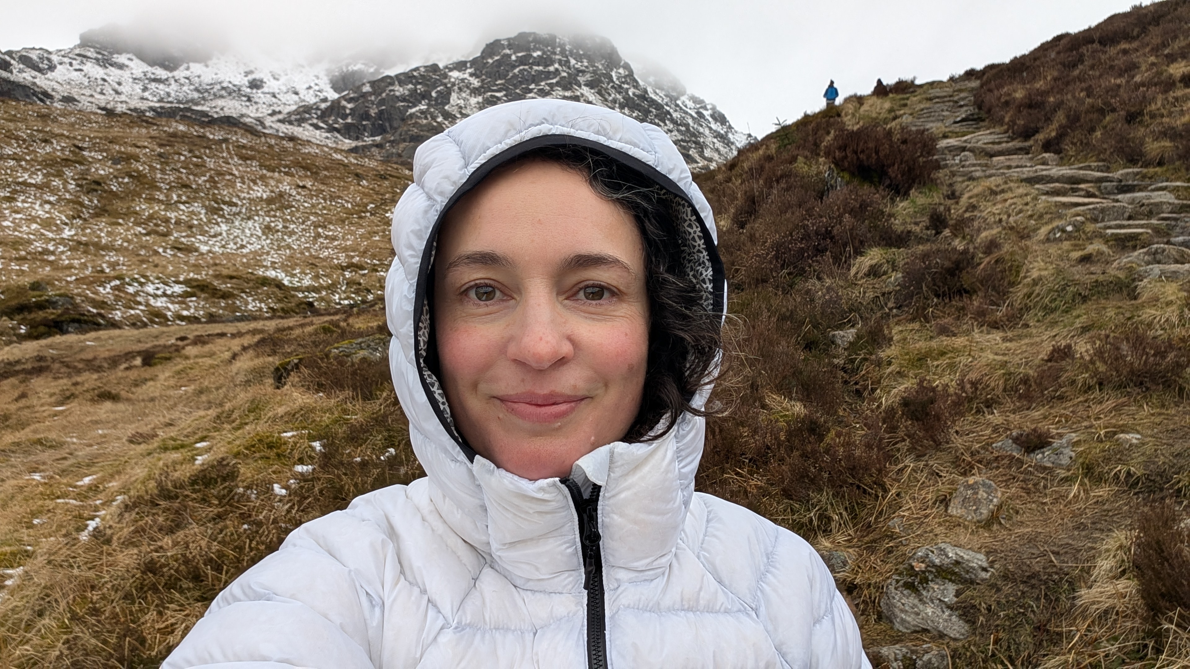 A woman takes a selfie while hiking in the mountains, wearing a white Columbia Arctic Crest Down jacket with the hood up