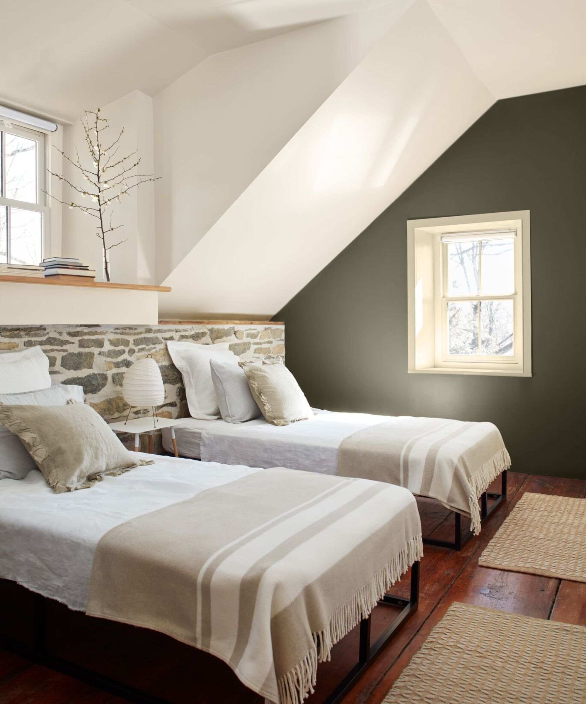 Twin bedroom with a wall painted in Cushing Green by Benjamin Moore, an exposed stone wall with two single beds draped with white and beige bedding in front, and wooden flooring with jute rugs.
