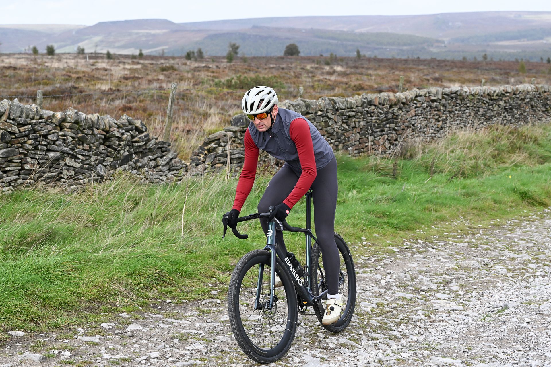 man riding a Green Basso Palta III gravel bike uphill out of the saddle on a limestone gravel track wearing a red jersey, grey gilet and grey tights