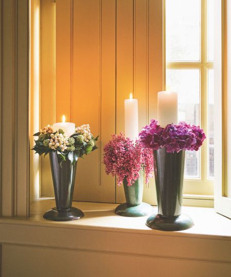 White pillar candles in vases filled with flowers, on a windowsill. There are yellow shutters behind, and a large window to the right.
